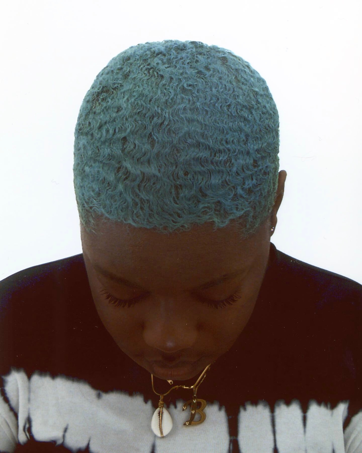 Self-portrait of artist Bernice Mulenga. They are looking down, their hair is blue, they are wearing a black and white top. You can see two chains around their neck, one has the letter be in gold and the other is of a cowrie shell.