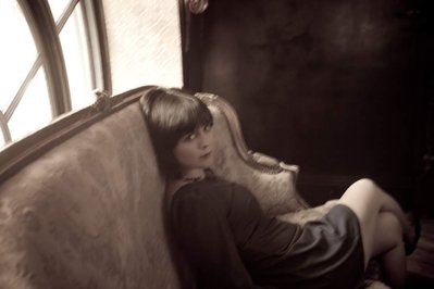 Vintage looking young caucasian woman with black hair and bangs and window behind her, lounges sensually on an antique couch