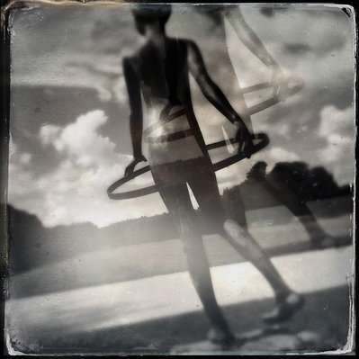 Black and white iPhone tintype of double exposure of young child hula-hooping in open green space on a cloudy day, shot from behind