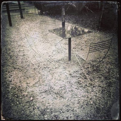 Black and white iPhone tintype of empty table and chairs surrounded by fallen leaves in park