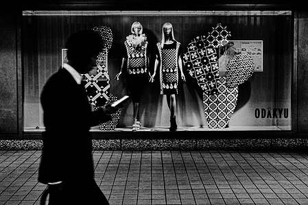 A figured gazes at their phone as they pass by the Odakyu department store, Tokyo. 
From the photographic series "In Praise of Shadows". Shot on black and white film by photographer Matthew Fleming. 