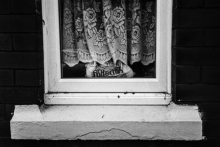 Photograph from the series To Live and Die in Lancashire: The Chapel of the Roses by Matthew James Fleming. Taken with a Leica M6 and Ilford HP5 35mm film. 
A bag of fish and chips in a window of a terraced house, Preston.