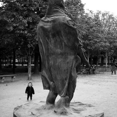 Black and white street photography of the french street photographer David Décamps representing a baby amazed in front of a giant statue in a park in Paris.