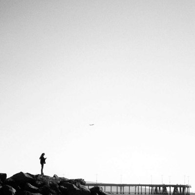Black and white street photography of the french street photographer David Décamps representing a woman on a rock with an airplane behind her in the beach of Los Angeles, USA.