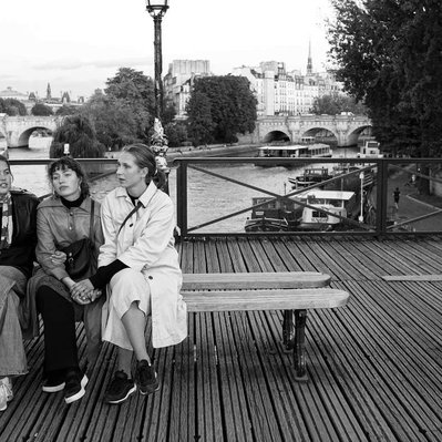 Black and white street photography of the french street photographer David Décamps representing three women sitting on a bench holding their hands on a bridge in Paris.