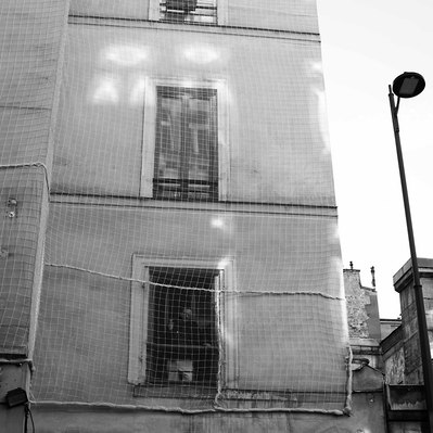 Black and white street photography of the french street photographer David Décamps representing a man at his window behind a site net in Paris.