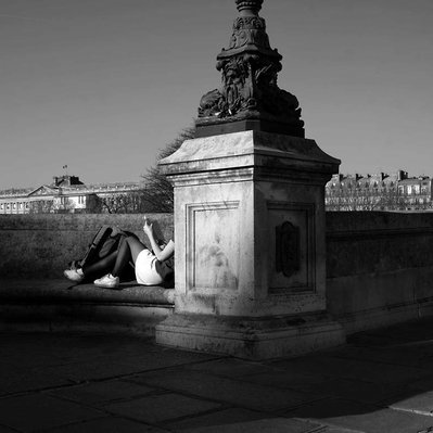 Black and white street photography of the french street photographer David Décamps representing a young girl reading a book with the sun on her legs in a bridge in Paris.