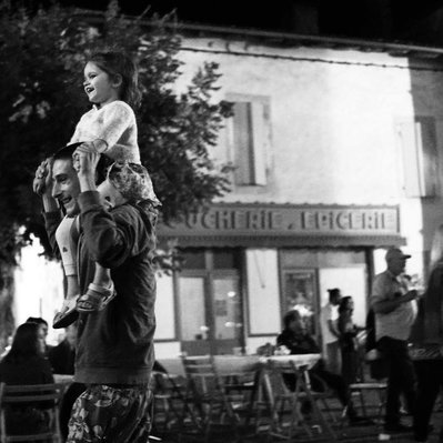 Black and white street photography of the french street photographer David Décamps representing a little girl on the shoulders of a man during a feast in a village.