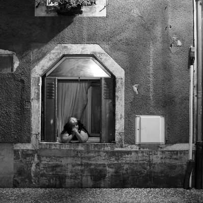 Black and white street photography of the french street photographer David Décamps representing a old woman alone at her window observing the village.