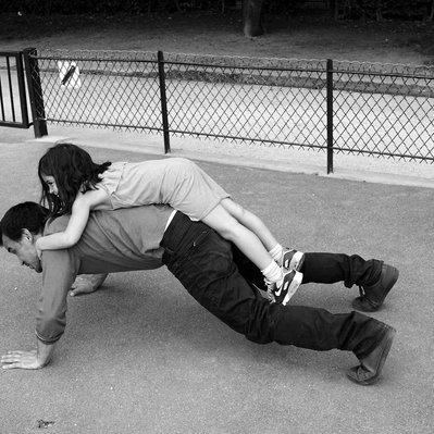 Black and white street photography of the french street photographer David Décamps representing a girl on the back of her father doing push up in a park in Paris.