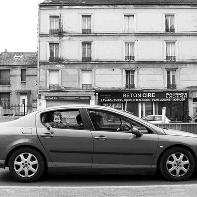 Black and white street photography of the french street photographer David Décamps representing a little boy doing a wince from a car in Paris.