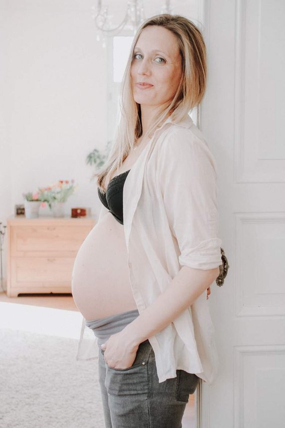 Schwangere Frau lehnt an Türrahmen mit offener Bluse so dass man ihren Babybauch sieht.