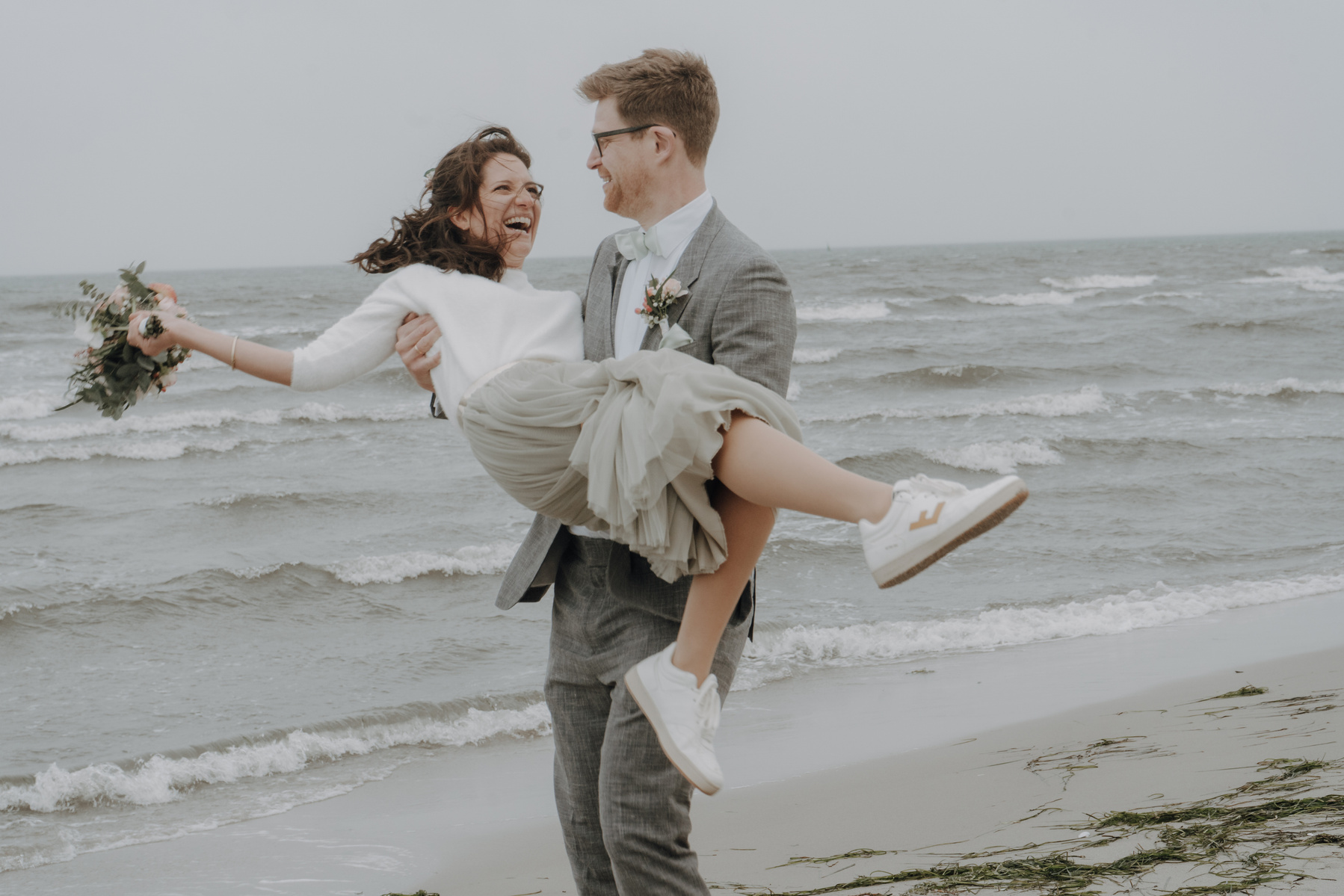 Bräutigam trägt Braut und wirbelt sie im Kreis mit Meer im Hintergrund.