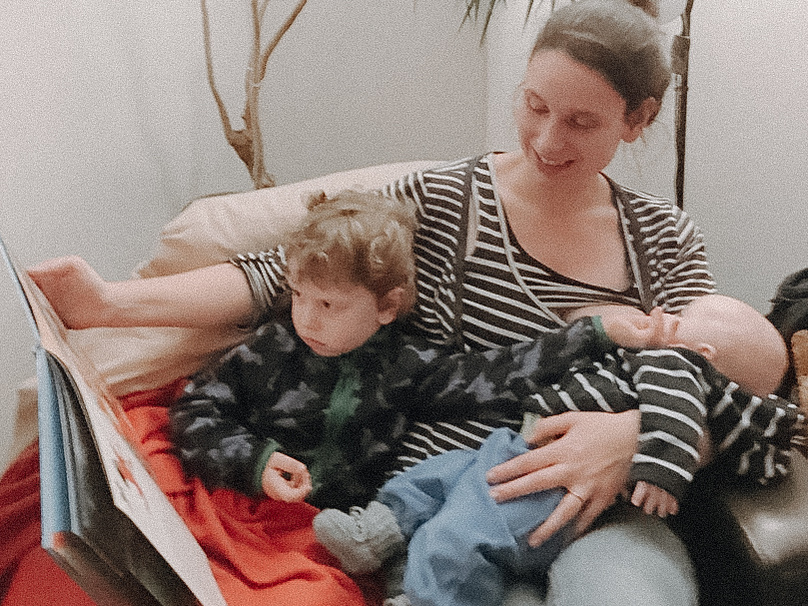 Junge Mutter stillt Baby und liest großem Geschwisterchen vor.