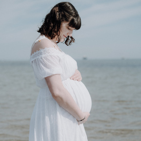Schwangere Frau am Meer im weißen Kleid schaut auf ihren Babybauch.