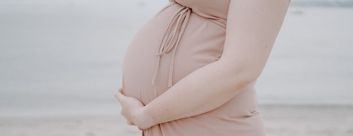 Nahaufnahme vom Babybauch im rosafarbenen Kleid mit Meer im Hintergrund.