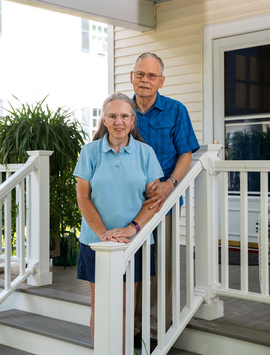 An older married couple stand on the steps of their porch in Vermont