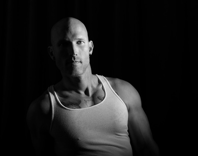 A man poses in a white tank top for a low key black and white portrait for The Man Project by Dark Light Portrait Studios of Williamsburg, VA