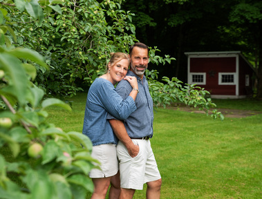 A married couple stand outside among apple trees