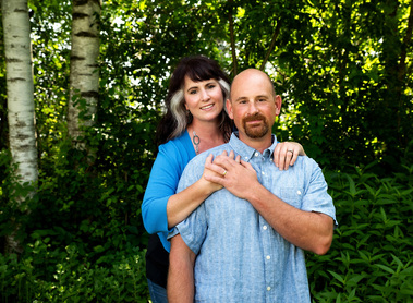 A married couple in blue shirts stand together outside at their home in Vermont