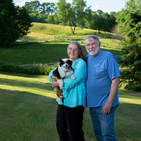 An older, married couple stand outside on their lawn with their dog in Vermont