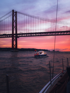 A small boat floats along the Tagus River under the 25 de Abril Bridge at sunset in Lisbon, Portugal. 