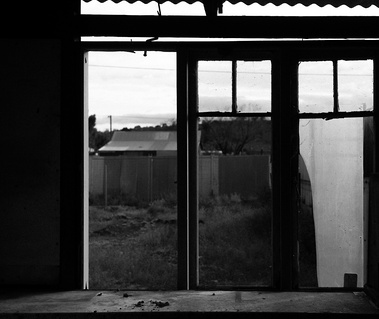 A black and white image that shows a view through an old window with broken glass, looking out into a backyard with a rooftop visible in the distance
