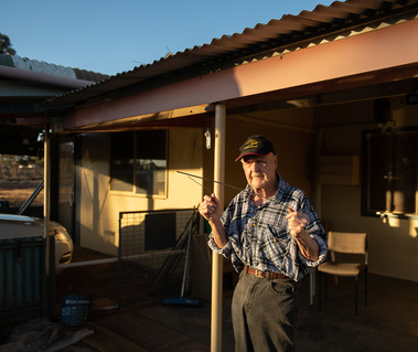 An older man holds two wires used for gold divining as he stands out the front of a house that is bathed in golden light. He is wearing a checked shirt, black jeans and a cap with KCGM on the front.