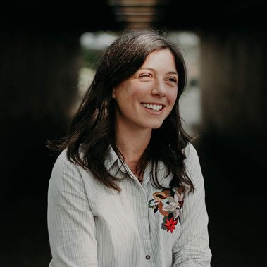 Photograph of Joanna Jowett, smiling white female, with long dark brown hair, sat with legs crossed and arms crossed at the wrist on her lap, wearing a white shirt with embroidery detail and burgundy trouser. Photo by Smart Banda.