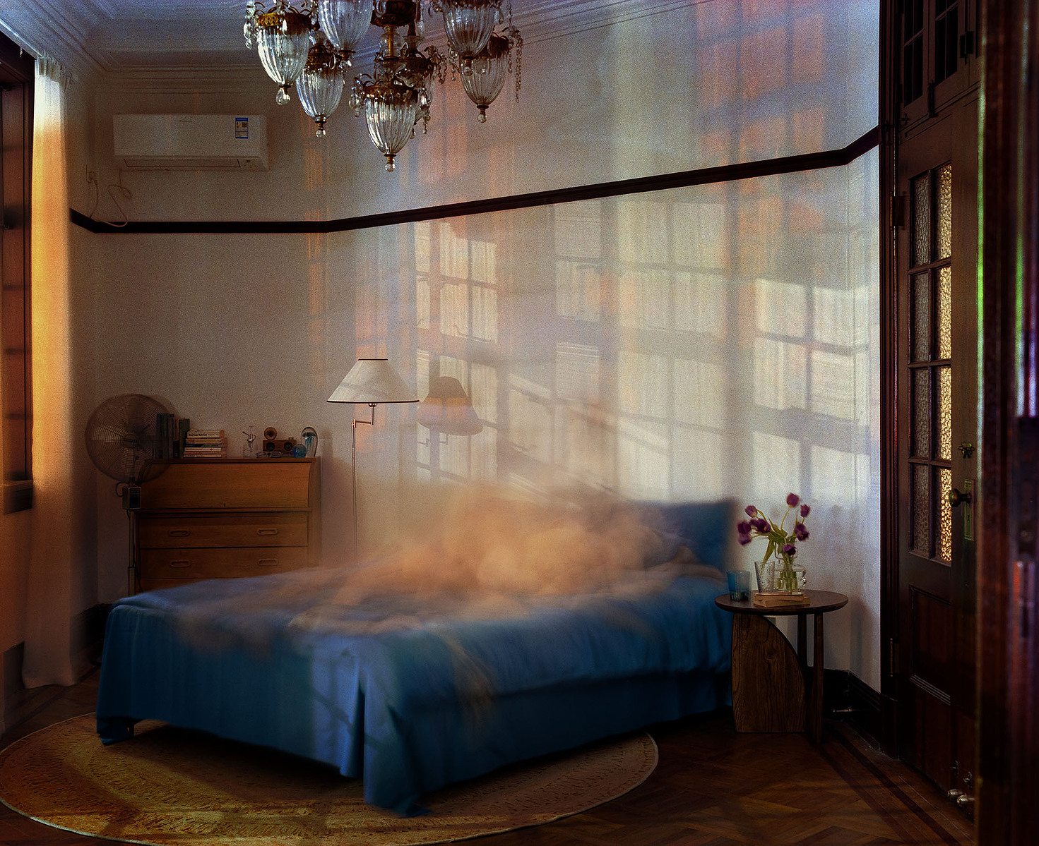 Blurred human figures on a blue bed atop a round carpet, with purple tulips and a blue glass on the bedside table. A standing lamp, fan, and a closet with books and decor are in the background, along with an old Shanghai-style door and ceiling lamp.