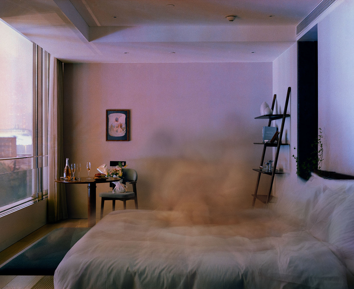 Blurred human figures on a white bed in the hotel, with wine, glasses, cake, and flowers on a wooden table. A small painting in the background, cityscape night view from the window, and a shelf with books and decor behind.