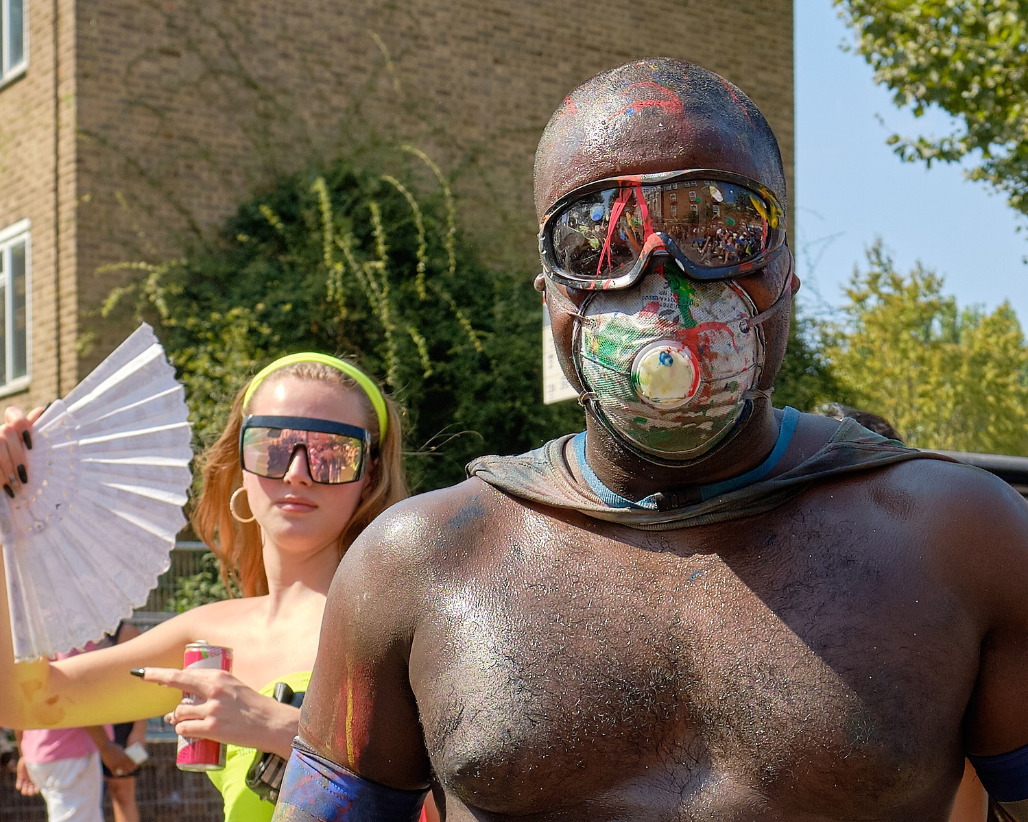 a topless man in a mask and a woman with a fan and sunglasses pose for a street portrait