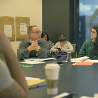 A group of people listening to a person in a grey sweater and glasses. 