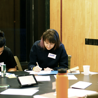 Two people looking down and writing on paper on the table and two people hugging next to them. 