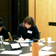 Two people looking down and writing on paper on the table and two people hugging next to them. 