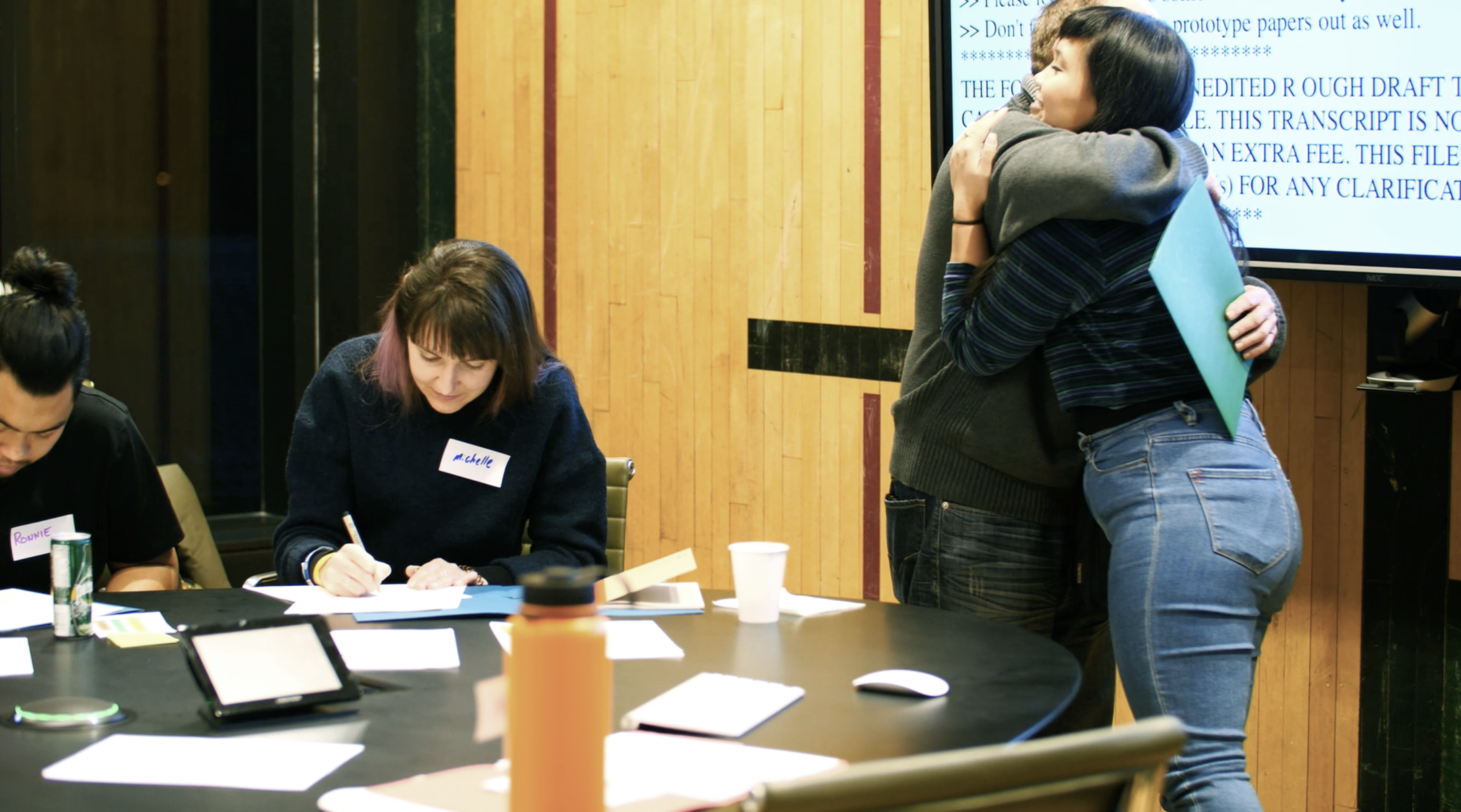 Two people looking down and writing on paper on the table and two people hugging next to them. 