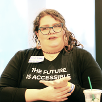 A person with curly hair, signing. They are wearing a shirt that says 'The Future is Accessible'.