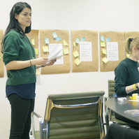 A person standing in front of a wall with sticky notes and people sitting at a table.
