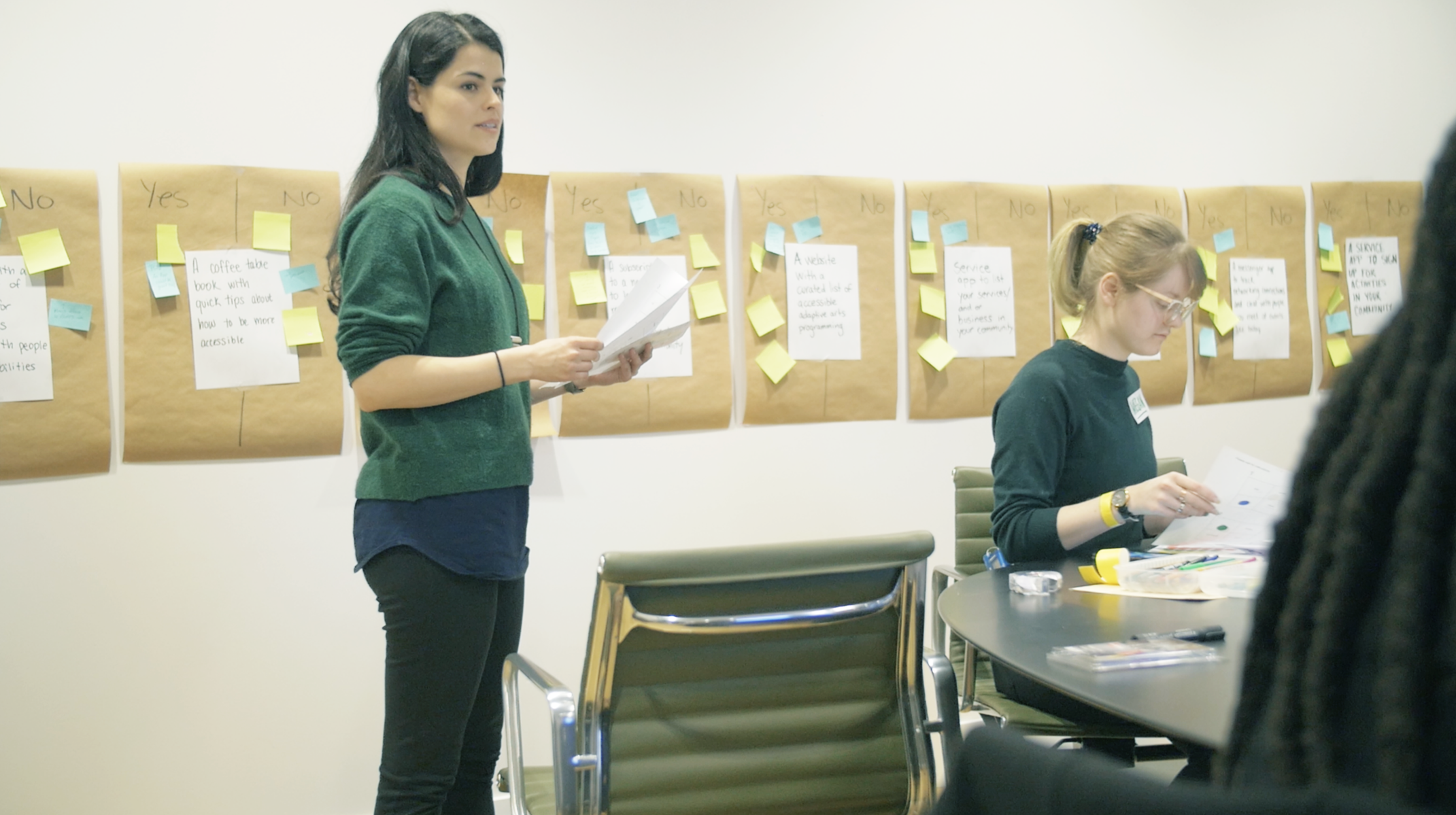 A person standing in front of a wall with sticky notes and people sitting at a table.