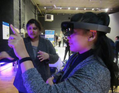 A person pointing wearing virtual reality goggles and another person showing them what to point at.