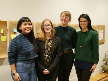 Four people smiling at the camera in front of a wall with paper and sticky notes.
