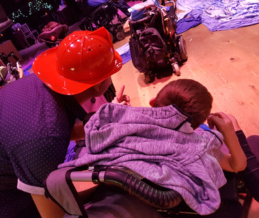 Support person wearing a firefighter hat next to person in a wheelchair at an event.