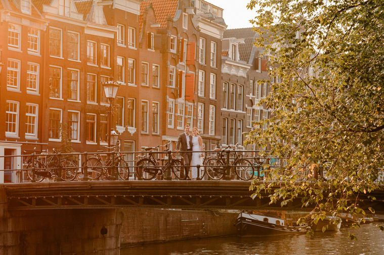 The bride and groom share a romantic moment on an Amsterdam bridge, surrounded by historic canal houses and bathed in the warm glow of sunset.