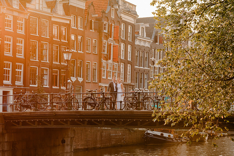 The bride and groom share a romantic moment on an Amsterdam bridge, surrounded by historic canal houses and bathed in the warm glow of sunset.