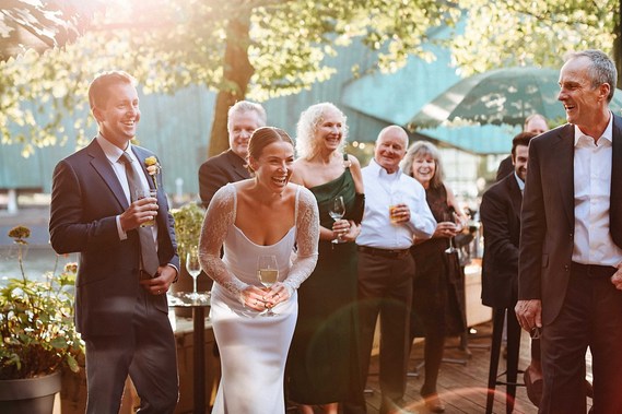 In warm golden hour light, the bride and groom share a joyous laugh with wedding guests, celebrating with drinks and smiles in a relaxed outdoor setting.
