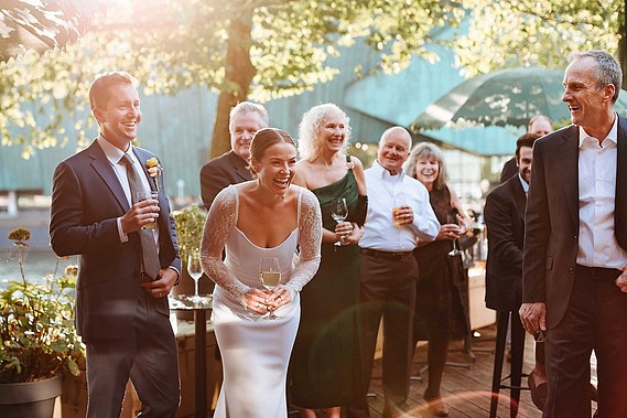 In warm golden hour light, the bride and groom share a joyous laugh with wedding guests, celebrating with drinks and smiles in a relaxed outdoor setting.