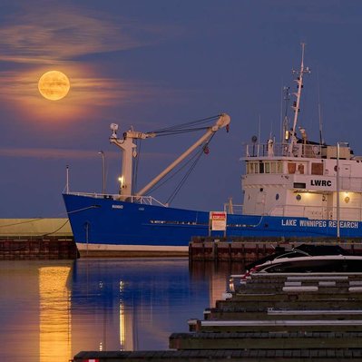 A full moon through wispy clouds rising above the Namao research vessel in Gimli harbour, Manitoba.
