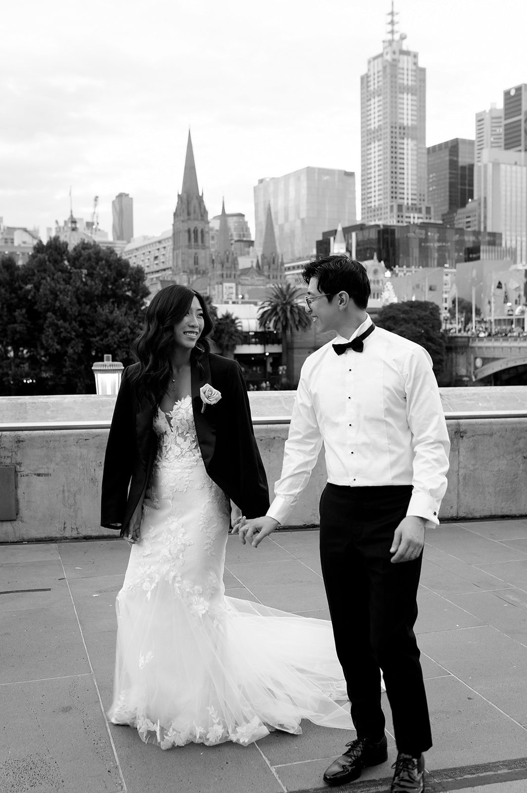 bride and groom walking in front of a cityscape. Bride has grooms black jacket on over her dress. 