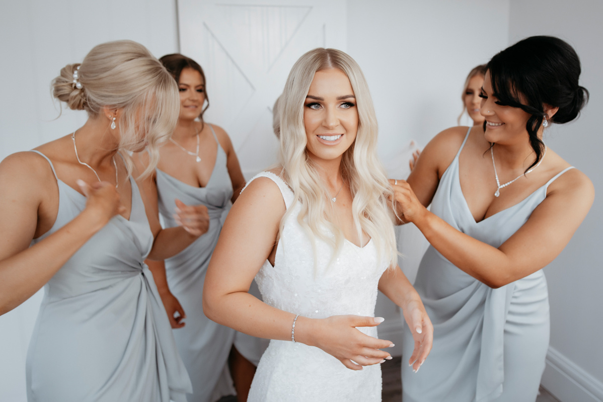4 bridesmaids, dressed in light grey dresses, helping the bride into an all-white wedding dress.