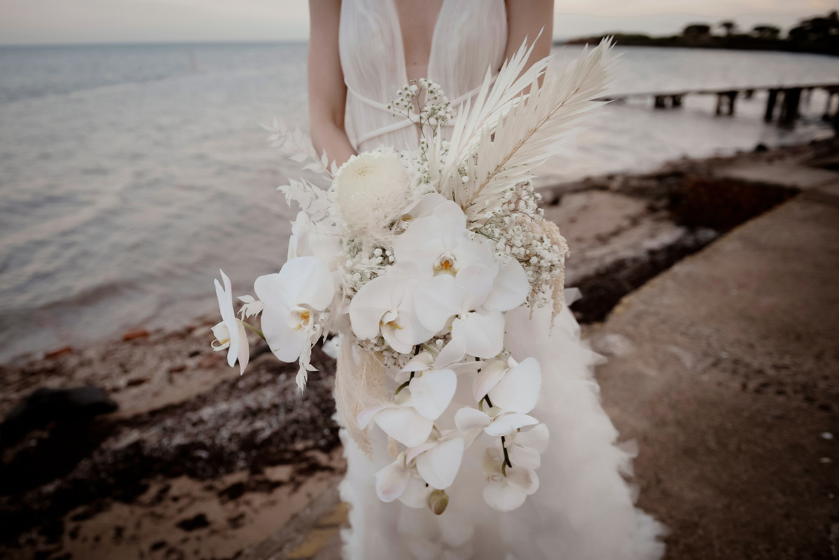 A close up picture of a white bouquet of flowers, held by a woman wearing a white wedding dress in front of the beach.